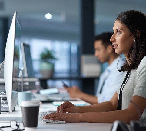 Businesswoman with a cup of coffee looks at her computer monitor while her coworker looks at his phone in the background